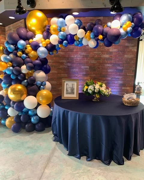 A navy, blue, gold, and white balloon arch framing a table with a navy tablecloth at a retirement party.