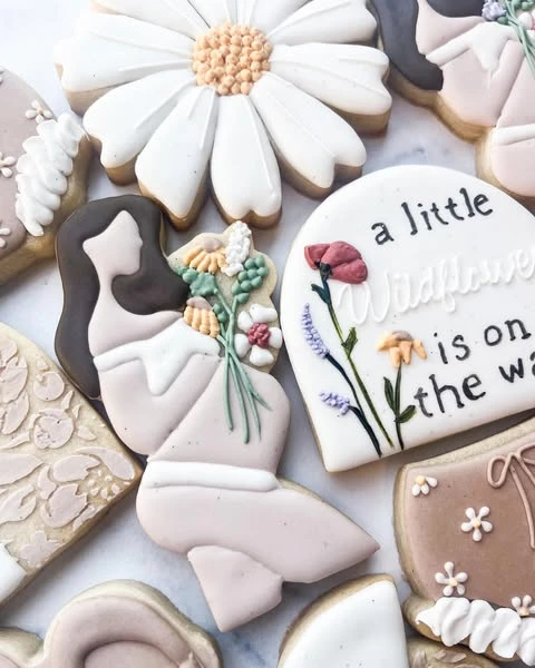 Custom baby shower cookie with the phrase 'a little wildflower is on the way' next to a cookie shaped like a mother holding a bouquet.