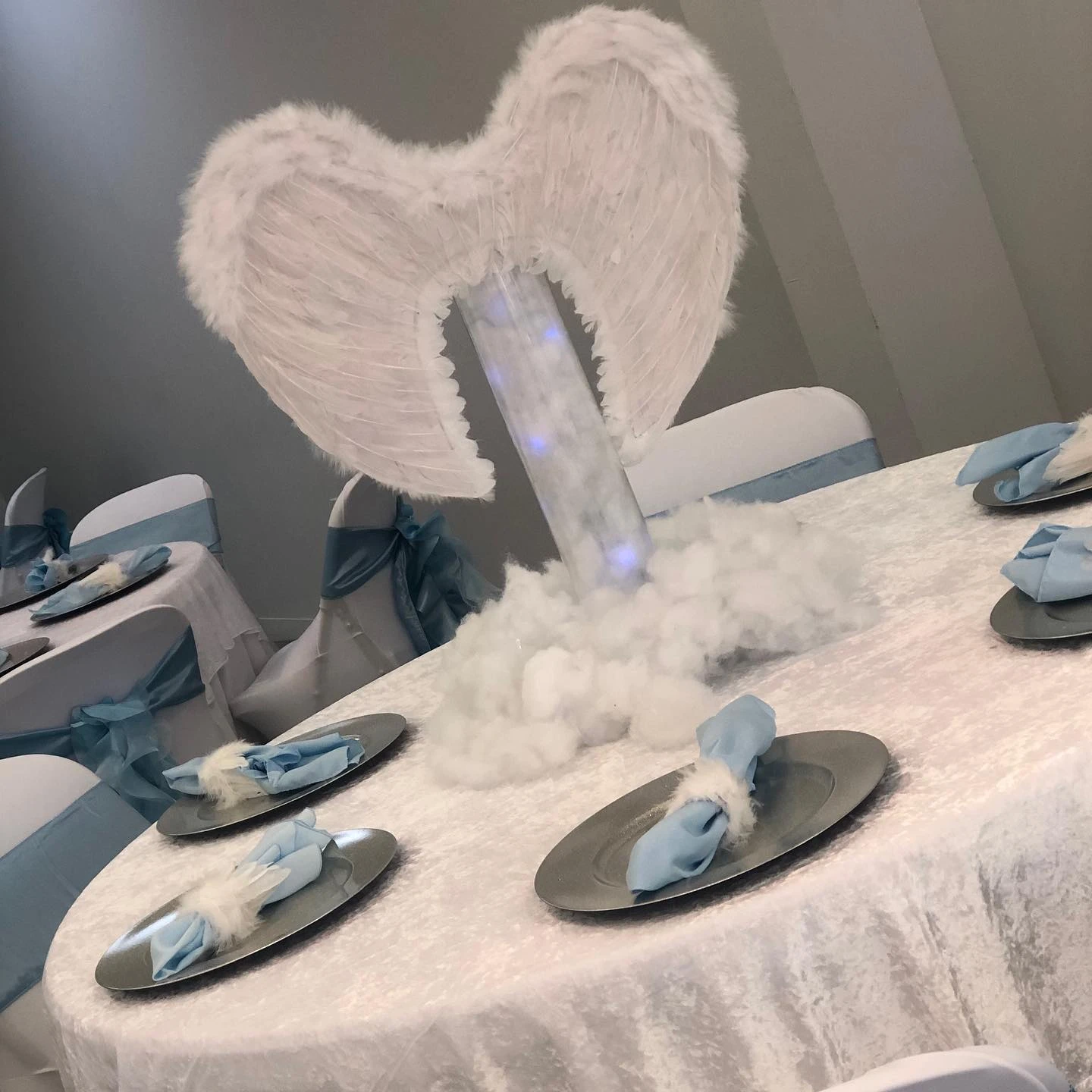 A tall 'Heaven Sent' baby shower centerpiece with white angel wings on an illuminated column rising from a fluffy white cloud base on a guest table.