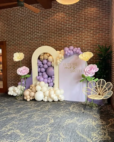 Spring bridal shower photo backdrop with a lavender and cream balloon garland set against arched panels and accented with giant paper flowers.
