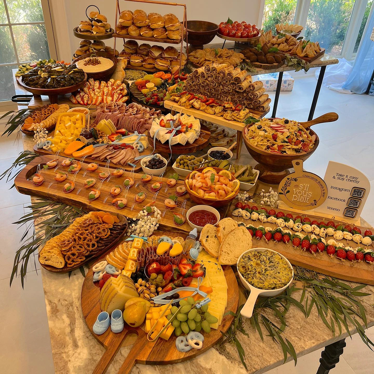 A lavish baby shower grazing table on a marble countertop, filled with charcuterie boards, tiered stands of sandwiches, and bowls of appetizers.