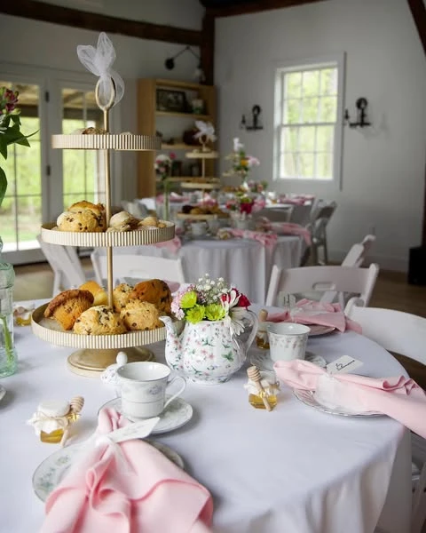 A tea party bridal shower table setting with a floral teapot full of flowers as the centerpiece, pink napkins, and a tiered stand with scones.
