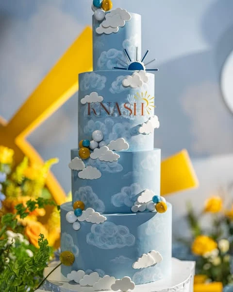 A four-tier blue baby shower cake decorated with white fondant clouds and a sun topper to match the 'Here Comes the Sun' theme.