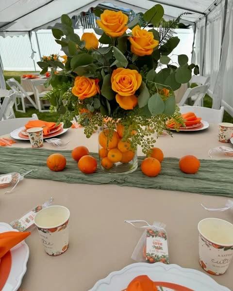 A baby shower table centerpiece in a clear glass vase filled with clementines, topped with an arrangement of orange roses and eucalyptus, set on a sage green runner.
