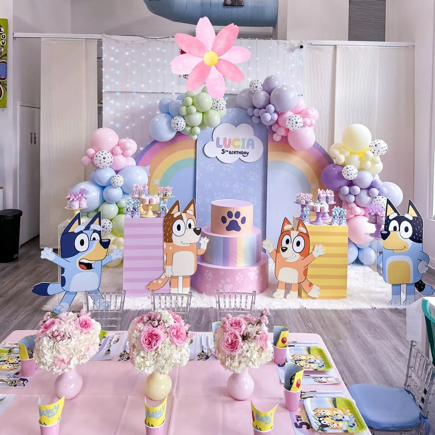 A Bluey birthday party dessert table featuring a large pastel rainbow backdrop, soft-colored balloon garlands, and cutouts of the Bluey characters.