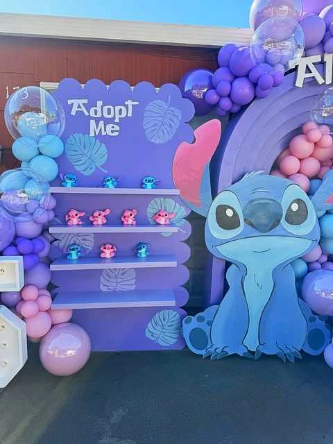 A purple 'Adopt Me' party favor station with shelves holding small Stitch and Angel plush toys for a birthday party.