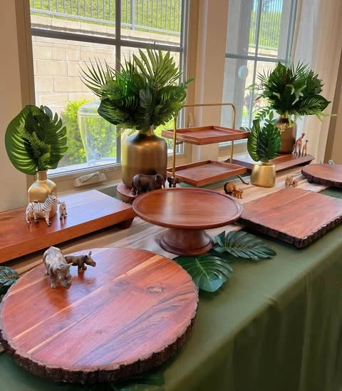Safari-themed baby shower food table setup with an assortment of empty wooden platters, tiered stands, and animal figurines on a green tablecloth.