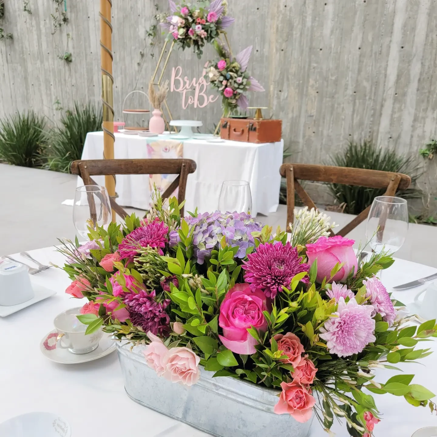Event table setting with a large floral centerpiece, wooden chairs, and vintage-style teacups.