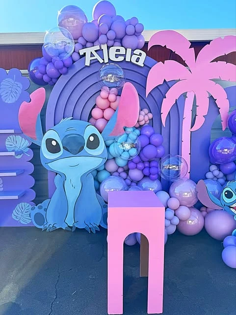 A Stitch birthday party backdrop with a large Stitch character cutout and a pastel balloon garland.