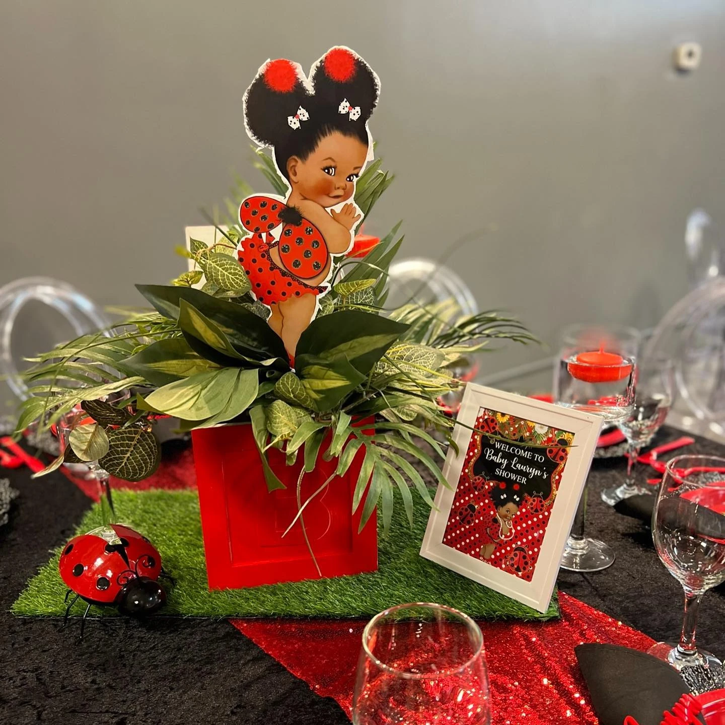 A ladybug-themed baby shower centerpiece featuring a baby character cutout in a red box of greenery, all set on a square of artificial grass.