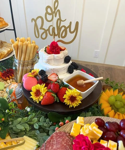 A of a two-tiered brie wheel cake on a dark stand, decorated with berries, flowers, and a gold 'Oh Baby' topper for a baby shower.
