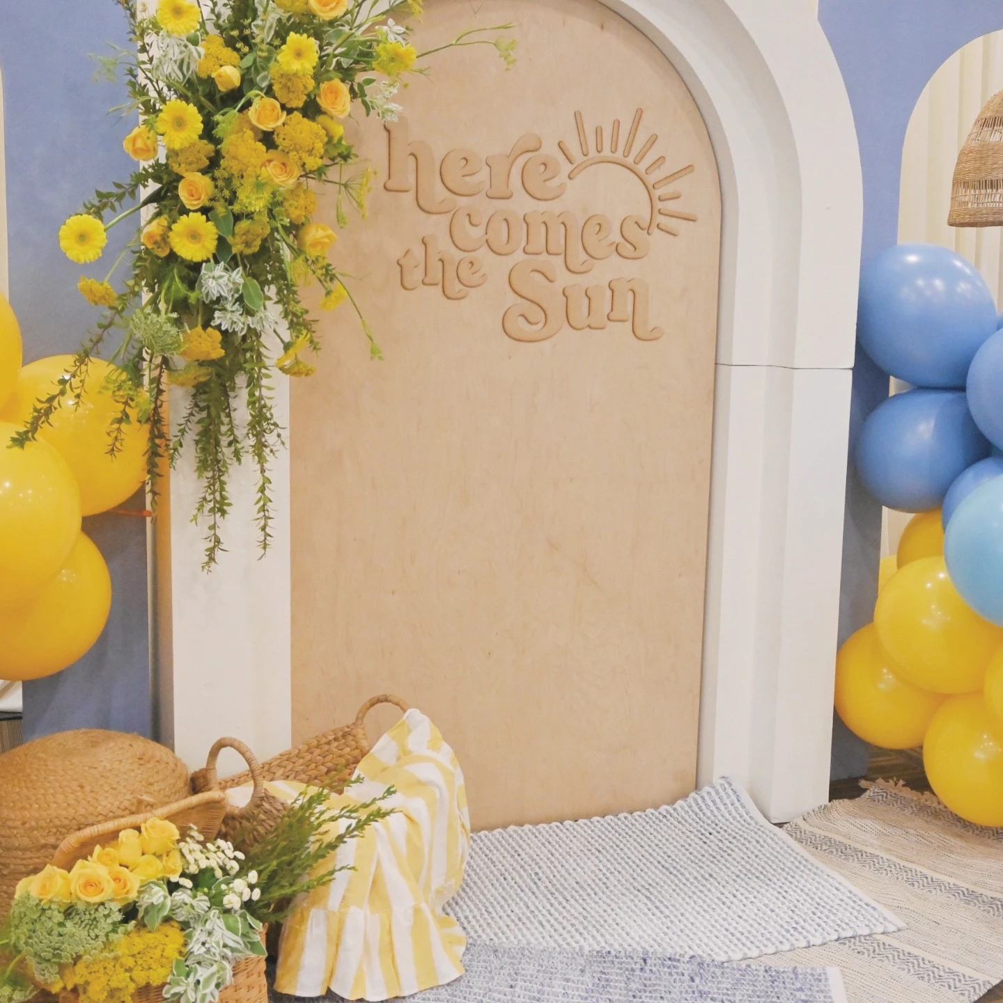 A 'Here Comes the Sun' baby shower backdrop featuring a wooden sign, yellow flowers, and blue and yellow balloons.