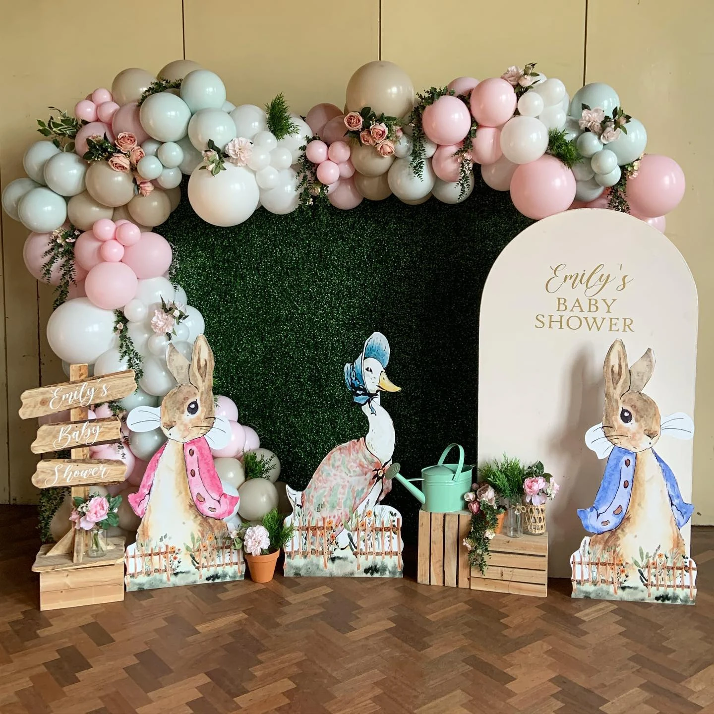A Peter Rabbit baby shower photo backdrop featuring a green hedge wall, a pastel balloon garland, and large cutouts of Peter Rabbit and Jemima Puddle-Duck.