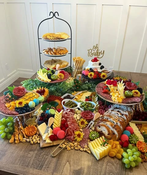 A wide view of a baby shower food table featuring a large grazing spread, a tiered stand, and a brie cake.
