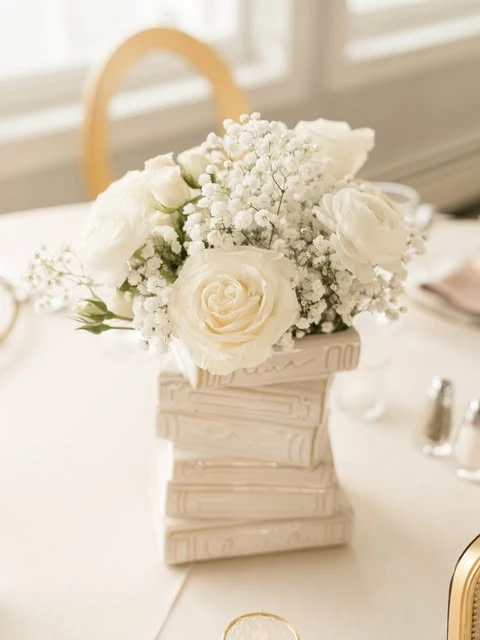 A baby shower centerpiece with white roses and baby's breath arranged in a vase shaped like a stack of white books.
