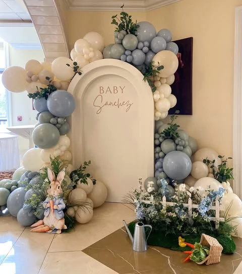 A Peter Rabbit baby shower photo backdrop featuring a blue and white balloon arch over a large character cutout and a small garden scene.