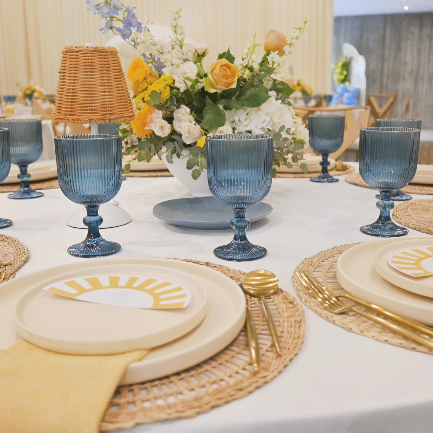 of a baby shower table setting with a sun-shaped place card, rattan charger, and blue glass.