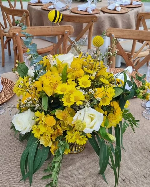 A rustic baby shower centerpiece with yellow flowers, white roses, and a bee accent in a gold vase on a burlap tablecloth.