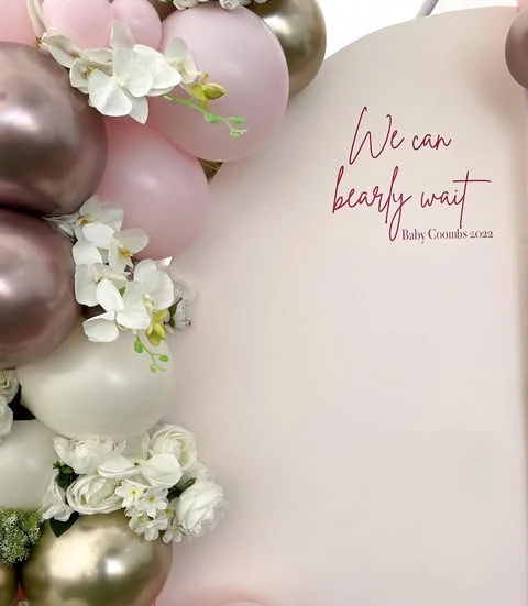 of the 'We can bearly wait' script on a pink backdrop with white flowers in the balloon garland.