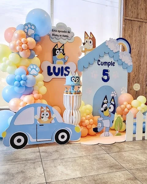 A full Bluey birthday party backdrop featuring a Heeler house cutout, character props, and a pastel balloon garland.