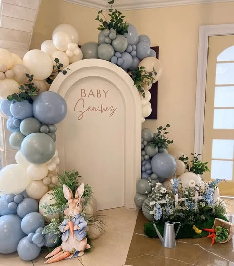 A of Peter Rabbit baby shower decor showing a miniature white picket fence, a metal watering can, and faux carrots on a patch of artificial turf.