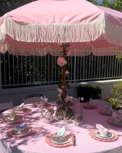 An outdoor tea party bridal shower table set under a pink umbrella with white fringe.