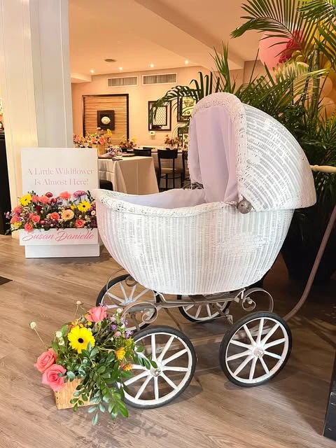 White vintage wicker pram next to a 'Little Wildflower' baby shower welcome sign.