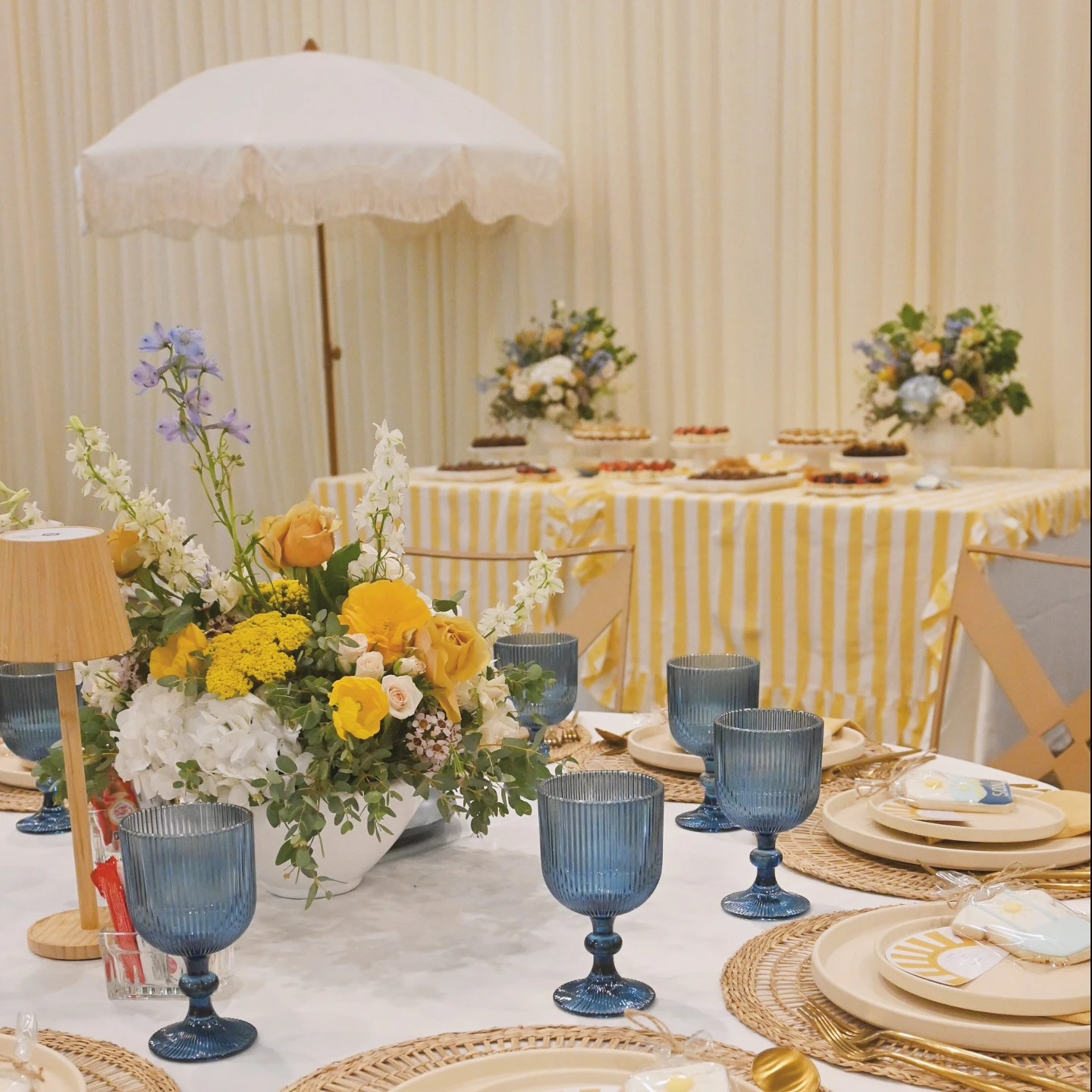 A 'Here Comes the Sun' baby shower table with a dessert station in the background featuring a striped tablecloth.