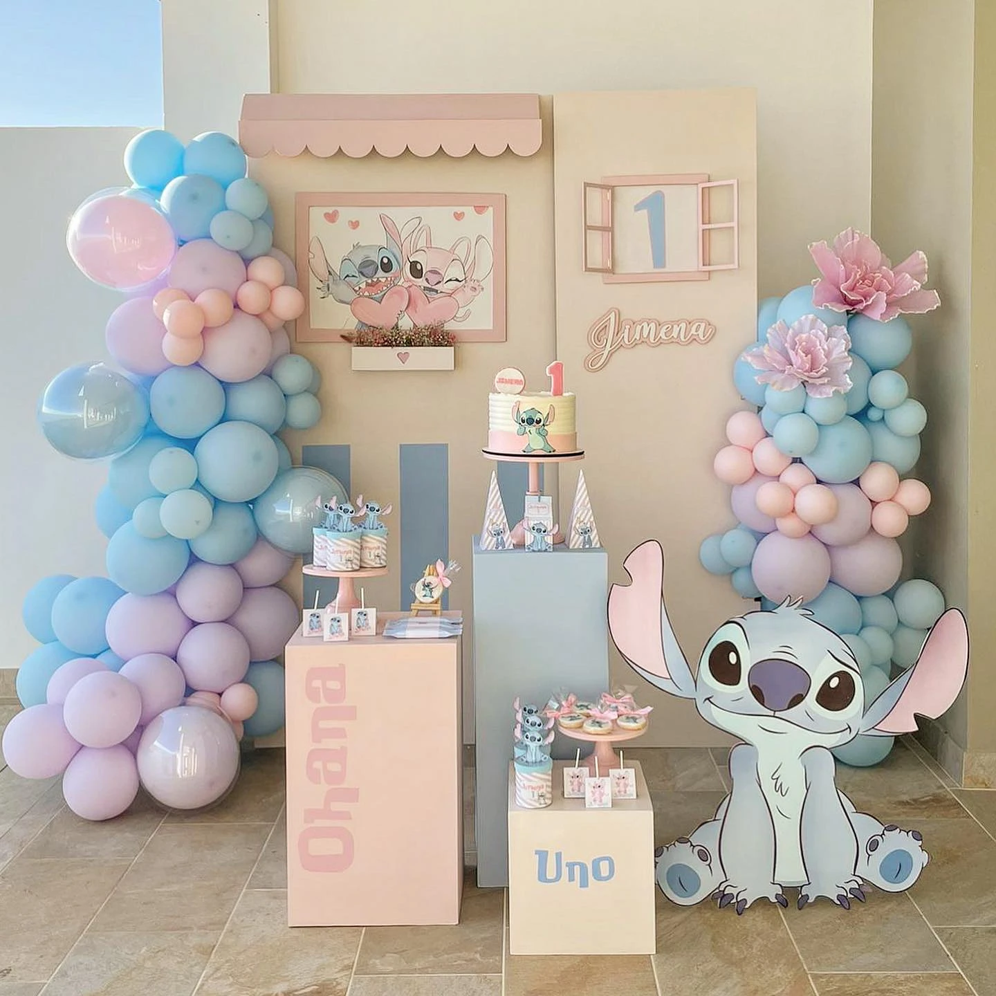 A Stitch-themed birthday party setup featuring pastel blue and pink balloon garlands, a large Stitch cutout, and a dessert display.