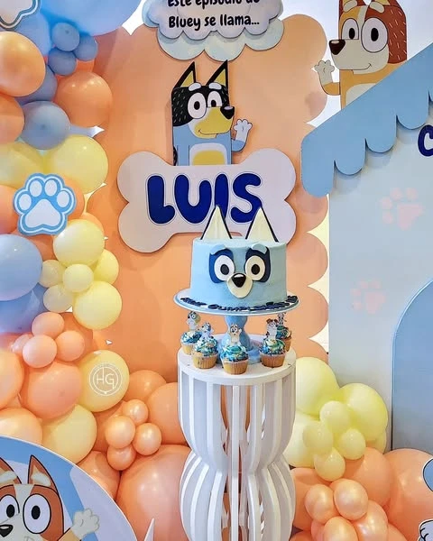 A Bluey-themed birthday cake and cupcakes on a white pedestal stand in front of a party backdrop.