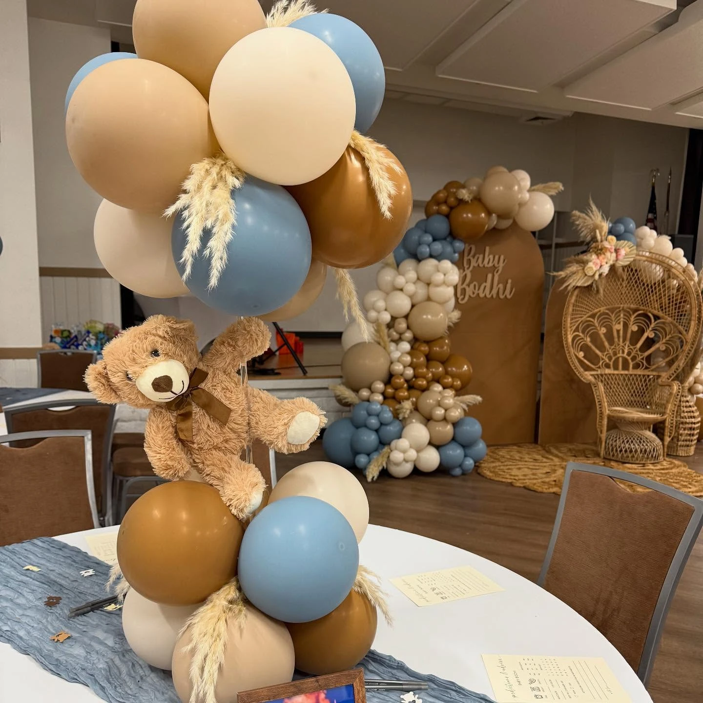 A 'we can barely wait' baby shower table centerpiece featuring a small teddy bear holding onto a cluster of balloons.