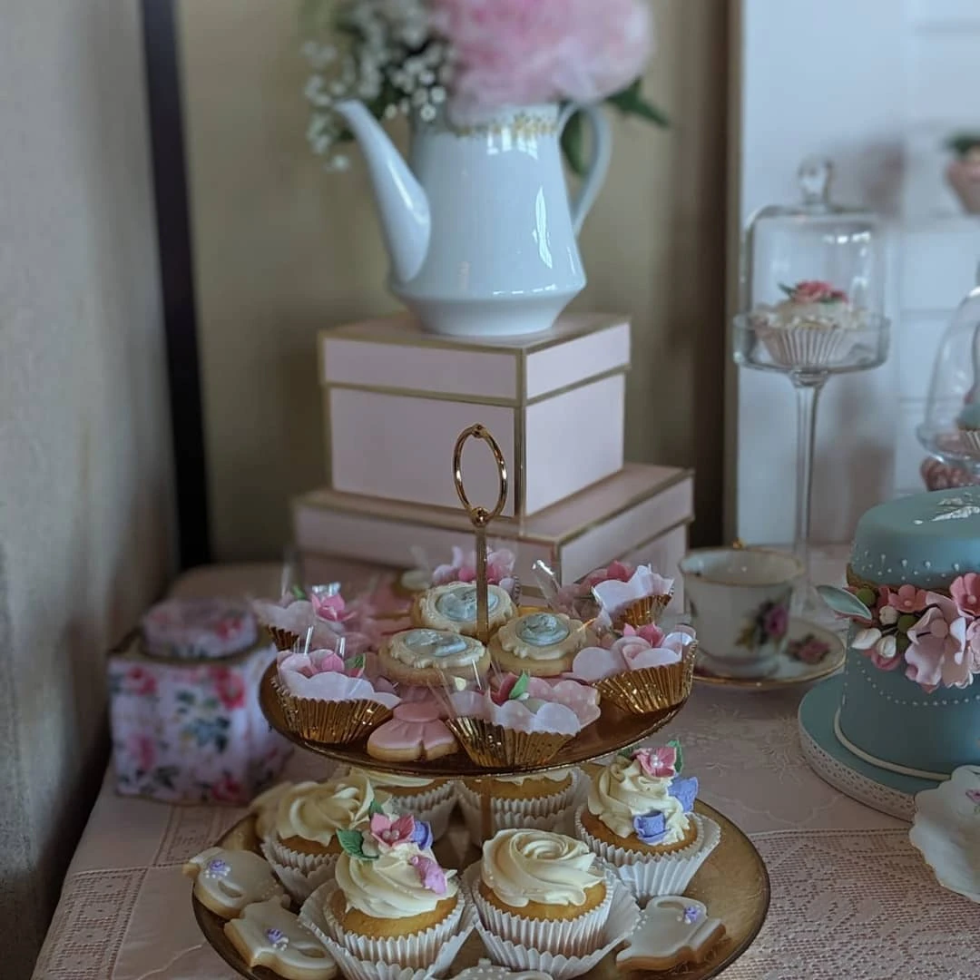 A two-tiered dessert stand with cupcakes next to a white teapot used as a flower vase for decor.