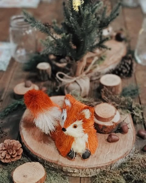 A woodland baby shower centerpiece with an orange fox figurine on a wood slice, surrounded by a mini pine tree, moss, and acorns.