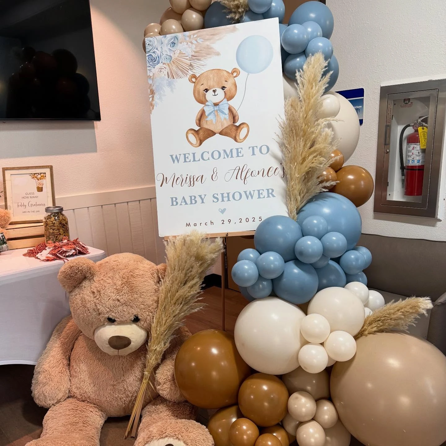 A welcome sign for a teddy bear baby shower, flanked by a large plush bear and a balloon arrangement.