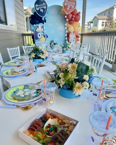 A long outdoor party table decorated for a Bluey birthday party, with a character backdrop and themed place settings.