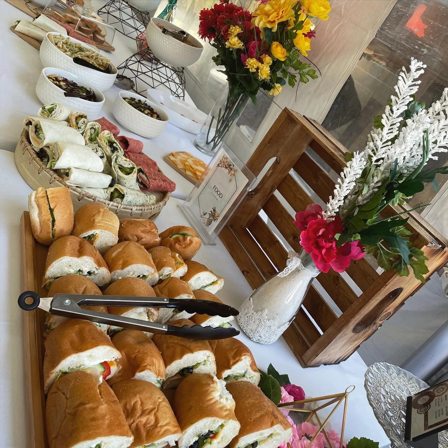 A baby shower food table setup with a variety of small sandwiches on a wooden tray and sliced tortilla wraps arranged in a woven basket.
