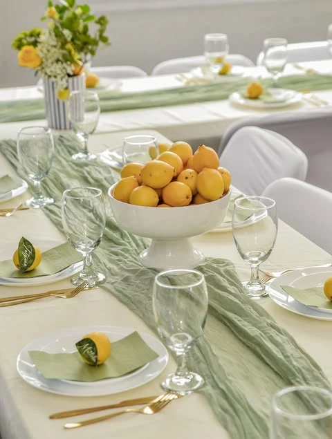 A baby shower table centerpiece featuring a white pedestal bowl overflowing with fresh yellow lemons on a sage green gauze runner.