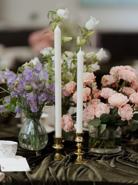 of pastel pink and purple flower arrangements next to white taper candles in brass holders on a velvet tablecloth.