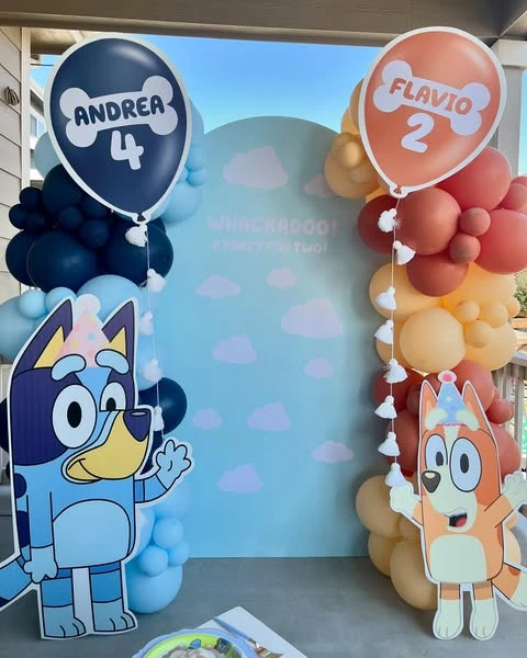 A Bluey and Bingo birthday party backdrop with character cutouts, balloon arches, and personalized bone-shaped signs.