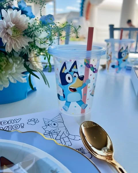 A reusable Bluey party cup with a lid and straw, next to a character coloring page placemat.