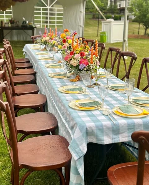Long guest table with a blue gingham tablecloth and colorful wildflower centerpieces for a baby shower.