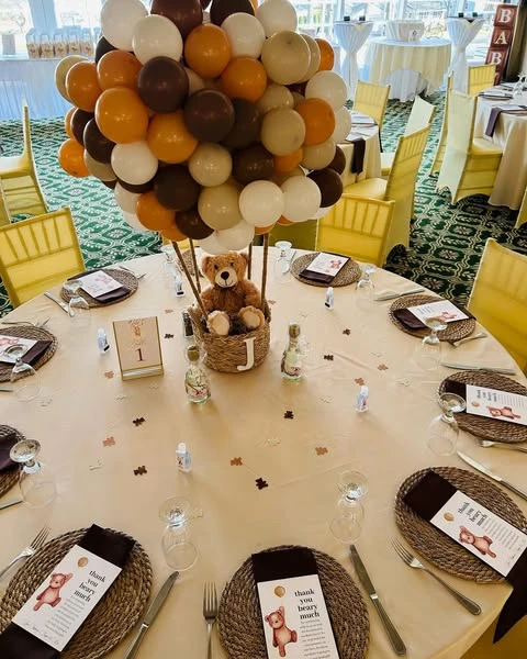 Teddy bear in a basket centerpiece under a cluster of brown and white balloons on a baby shower table.