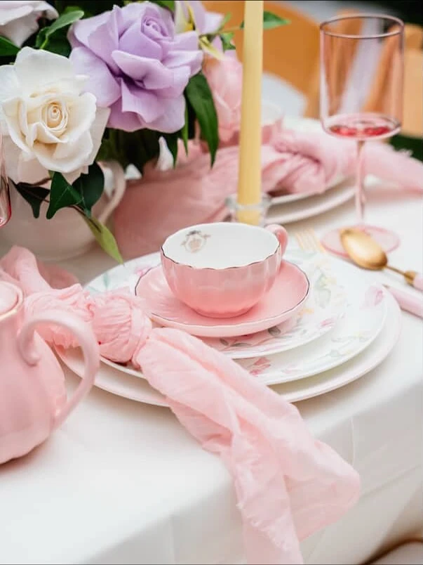 A tea party place setting with a pink teacup, floral plate, gold spoon, and a loosely tied pink napkin.