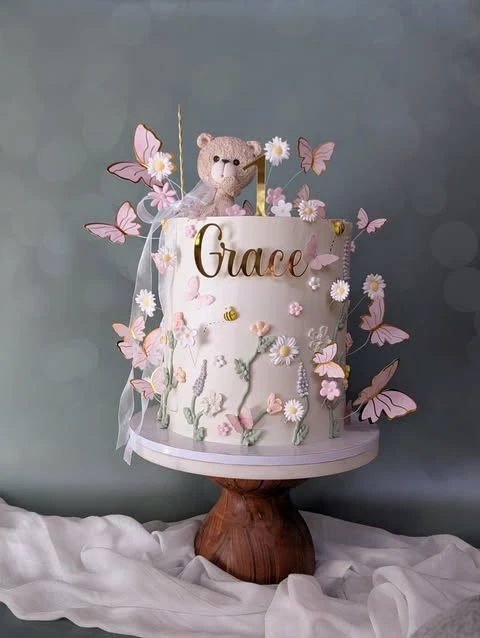 A tall wildflower-themed baby shower cake with pastel flowers, pink butterflies, and a teddy bear topper.