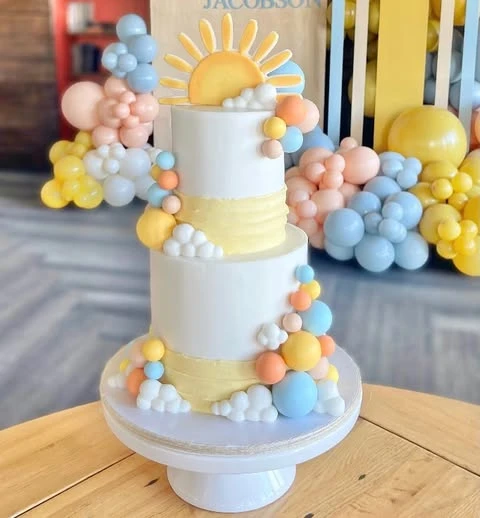 A two-tiered 'here comes the sun' baby shower cake with a sun topper and pastel decorations.
