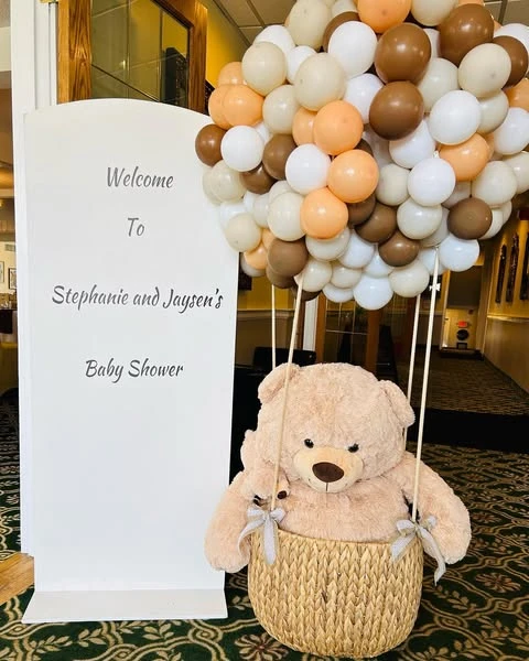 Baby shower welcome sign next to a large teddy bear in a basket with a brown and white balloon garland.