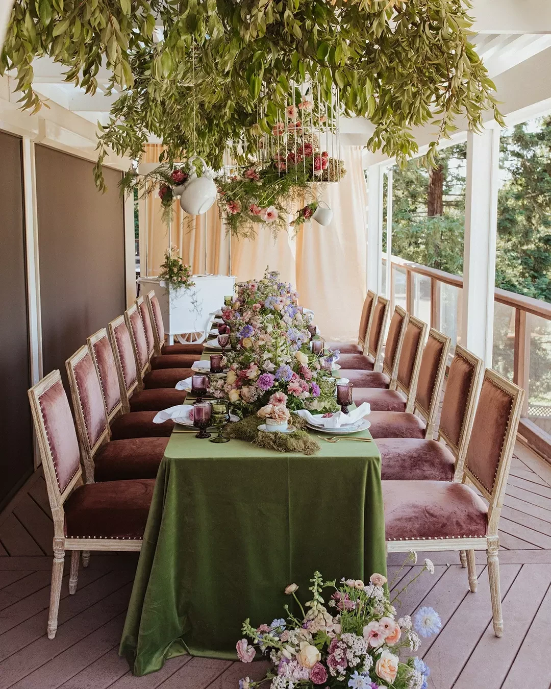 A long dining table set for a garden tea party bridal shower with a lush hanging installation of greenery and white teapots.
