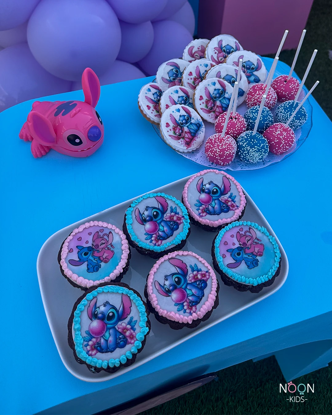 Plates of Stitch and Angel themed cupcakes, cookies, and cake pops on a blue table.
