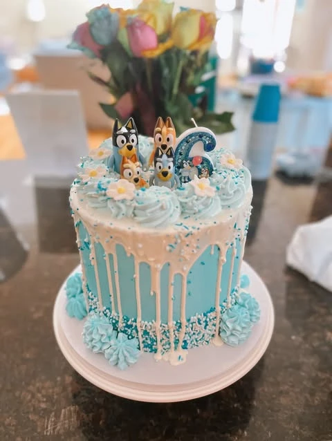 A light blue Bluey birthday cake with white drip icing and the Heeler family figurines on top.