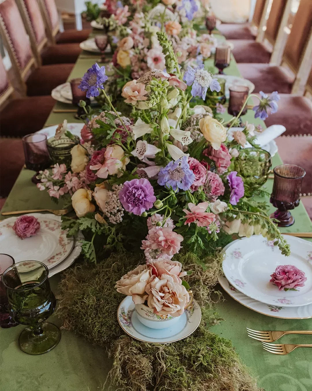 A tea party place setting with a teacup used as a small flower vase on a moss table runner.